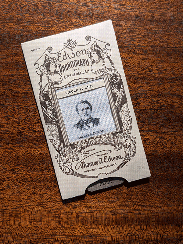 A photograph of a handheld novelty with a pull tab adertising the Edison Phonograph